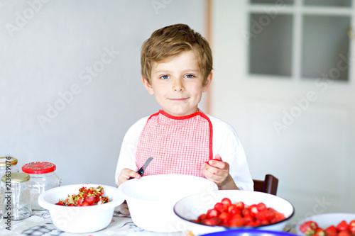 Little blond kid boy helping and making strawberry jam in summer