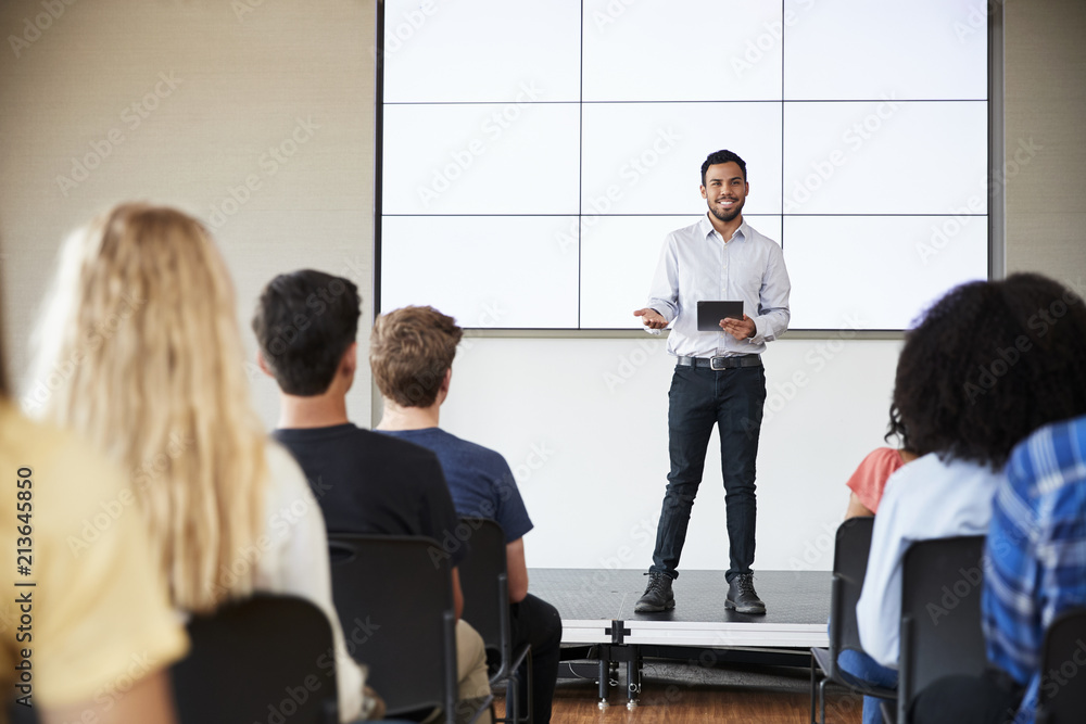 Male Teacher With Digital Tablet Giving Presentation To High School Class In Front Of Screen