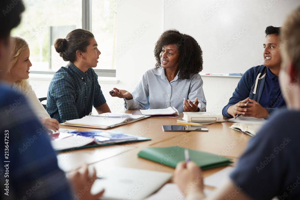 Female High School Tutor Sitting At Table With Pupils Teaching Maths Class Female High School Tutor Sitting At Table With Pupils Teaching Maths Class