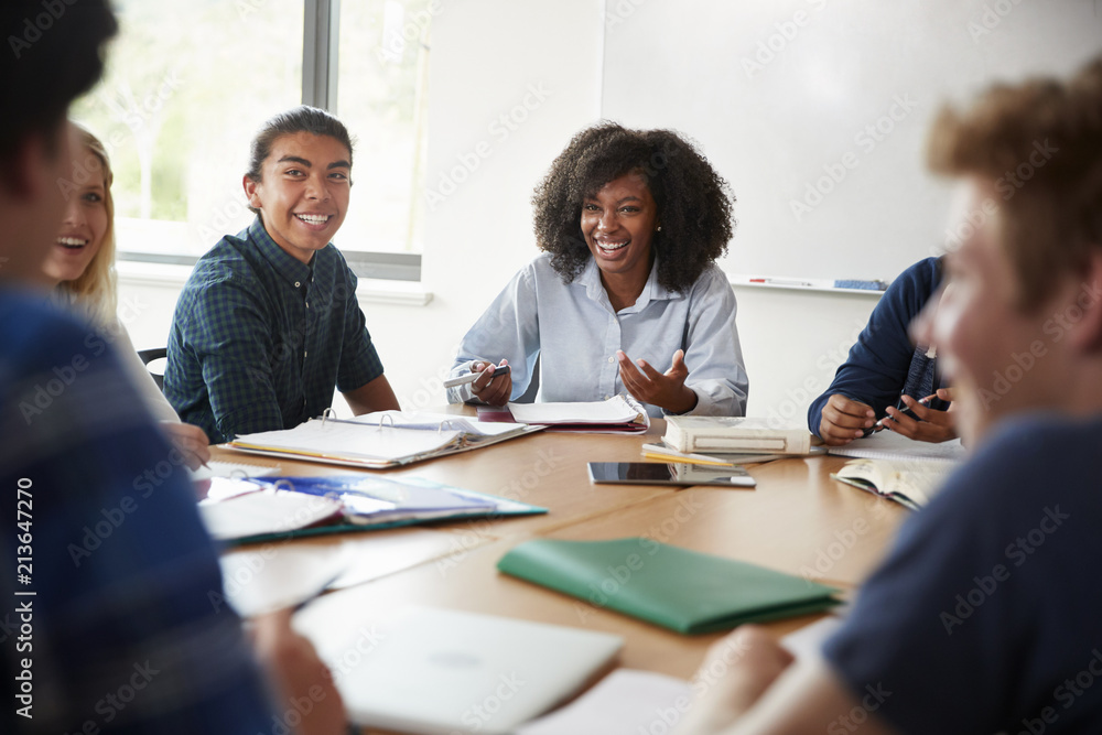 Female High School Tutor Sitting At Table With Pupils Teaching Maths Class Female High School Tutor Sitting At Table With Pupils Teaching Maths Class