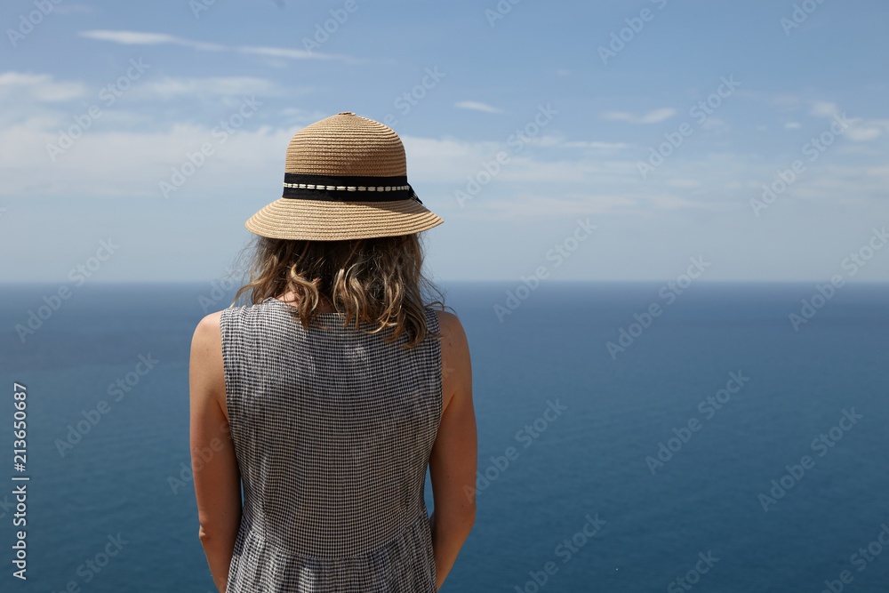 Closeup back view of unrecognizable slender blonde woman in dress and straw hat enjoying amazing seascape at viewpoint. Romantic female admiring picturesque scenery over vast calm ocean and blue sky