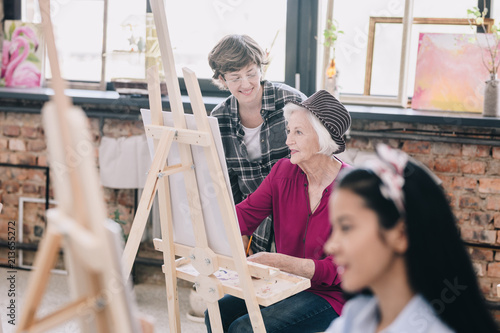 Portrait of modern senior woman painting sitting at easel in art studio studying art with smiling female teacher giving comments, scene in spacious sunlit loft space.