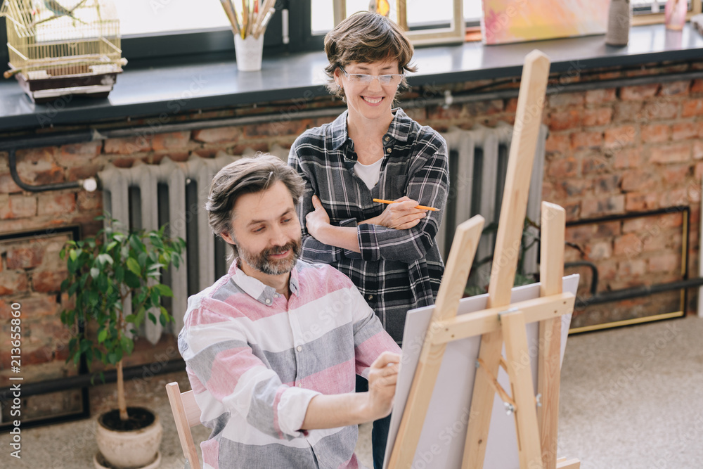 Portrait of smiling mature man painting sitting by easel in art studio with female art teacher watching him in art class, copy space
