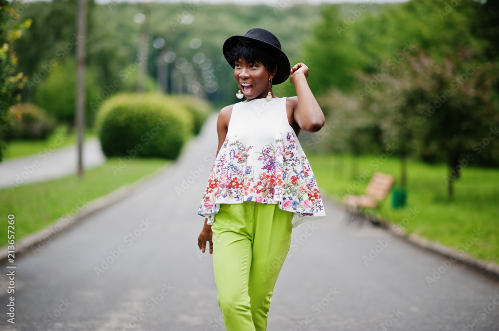 Amazing african american model woman in green pants and black hat posed with different emotions at park.