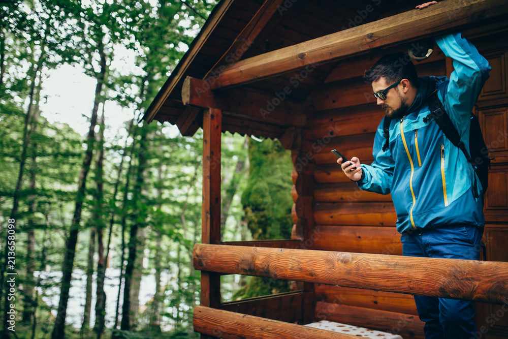 Man standing at the balcony of wooden house in forest and using mobile phone Man standing at the balcony of wooden house in forest and using mobile phone