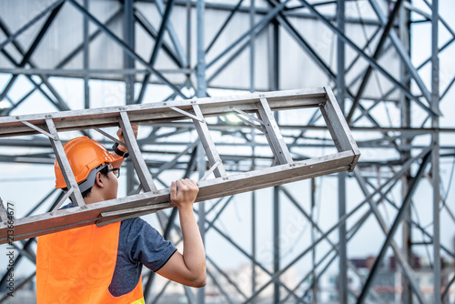 Young Asian maintenance worker with orange safety helmet and vest carrying aluminium step ladder at construction site. Civil engineering, Architecture builder and building service concepts