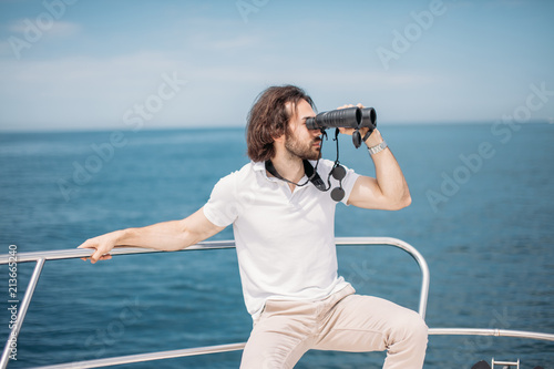 European bearded turist, having a boattrip, looks through binoculars from the boat, sitting at bow of yacht, looking at sea expecting to see dolphins. photo