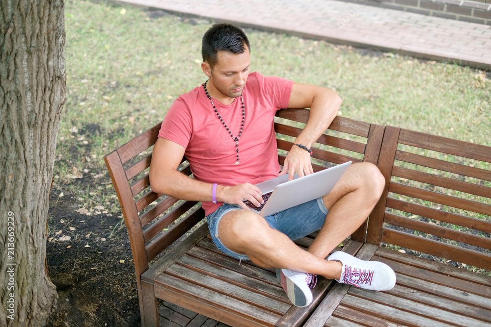 Молодой, красивый, здоровый, мужчина, сидит на скамейке с ноутбуком и набирает новый пост в блог
