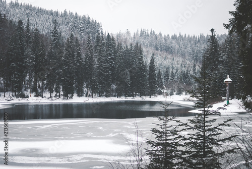 Beautiful mountain lake Synevyr in Ukraine. Frozen lake covered with snow against dark winter forest and mountains covered with clouds. Grey cloudy sky
