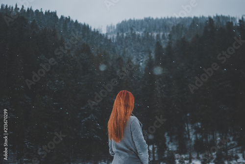 Handsome redhead girl looking in the distance. Splendid shady black mountain lake reflecting trees of forest behind her. Dreamy winter landscape of Carpathian mountains