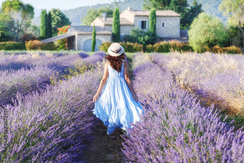 Fascinating young French girl walking the field of lavender in Provence, France, national park Luberon. Fashion outfit blue dress, straw hat. Back view. Traditional house in background.