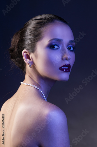 Εκτύπωση καμβά Beautiful brunette girl with naked shoulders and dark red lips make-up, wearing earrings and a pearl necklace, illuminated in blue