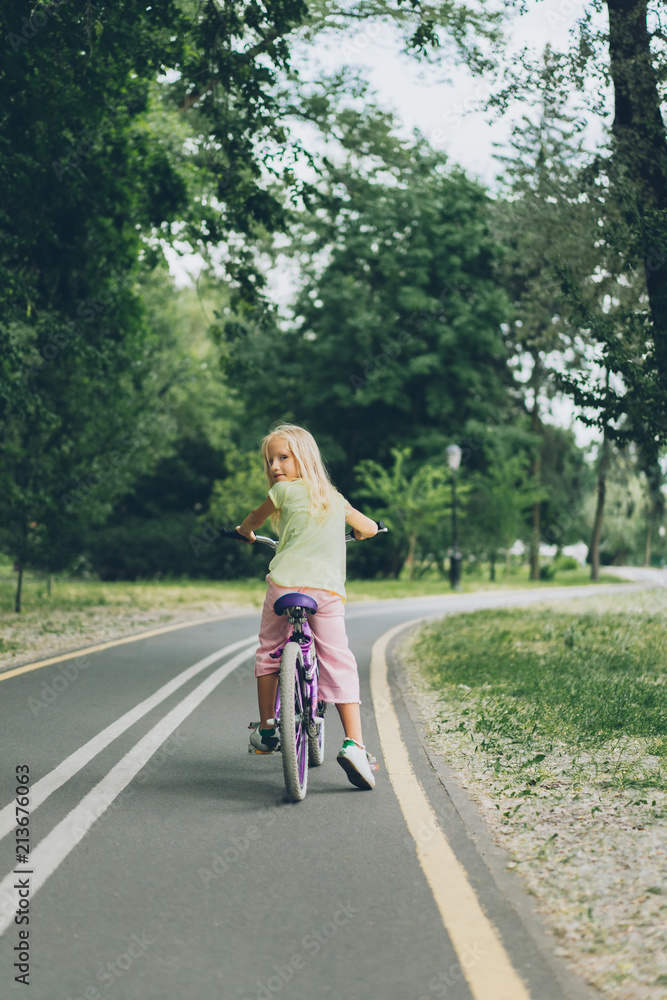 back view of kid looking at camera while riding bicycle on road in park back view of kid looking at camera while riding bicycle on road in park