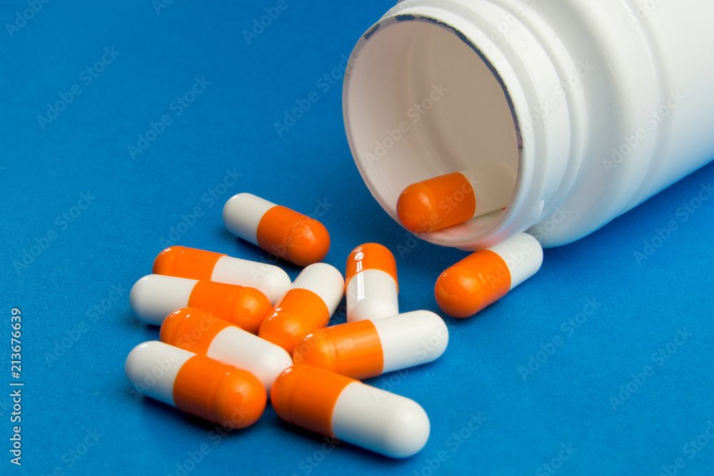 Orange white capsules (pills) were poured from a white bottle on a blue background. Medical background, template. Orange white capsules (pills) were poured from a white bottle on a blue background. Medical background, template.