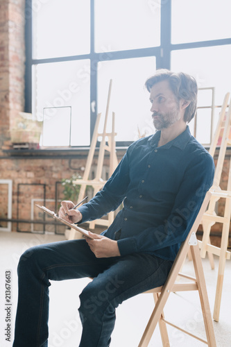 Warm toned full length portrait of pensive mature man sitting alone in art studio and drawing sketches