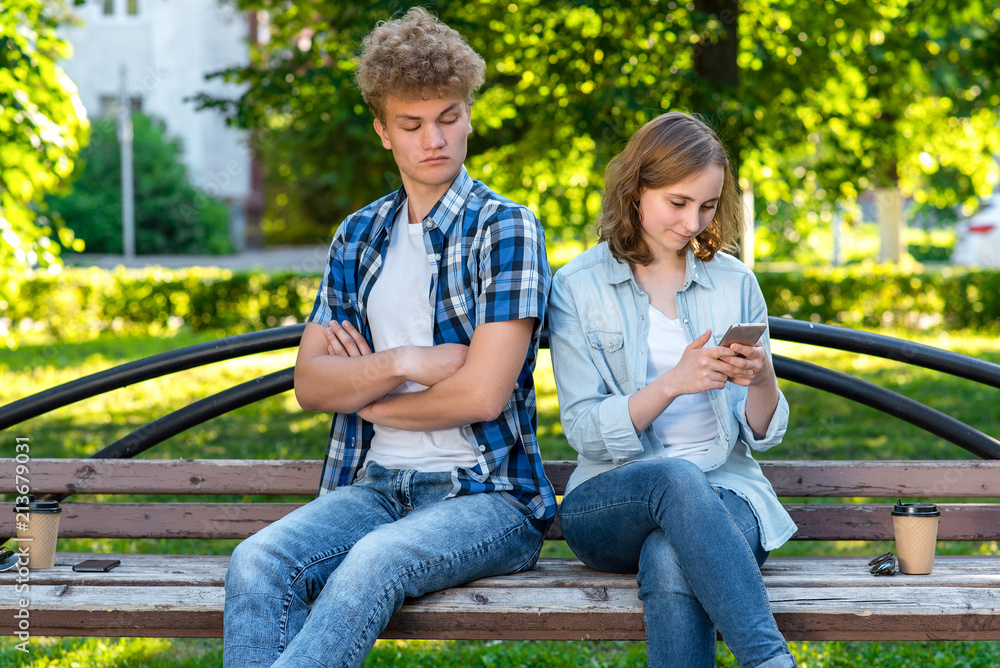 The guy and the girl are sitting on bench. The girl writes messages in social networks. The guy spies on correspondence. The concept of distrust in a relationship. Family problems. The guy and the girl are sitting on bench. The girl writes messages in social networks. The guy spies on correspondence. The concept of distrust in a relationship. Family problems.