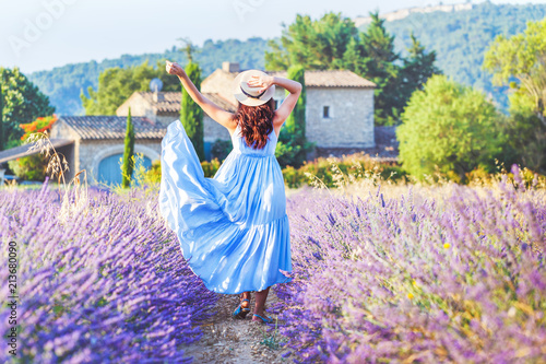 Beautiful young woman enjoy blooming lavender in Provence, France, national park Luberon. Fashion outfit blue dress, straw hat. Back view. Traditional house in background. Violet in nature.