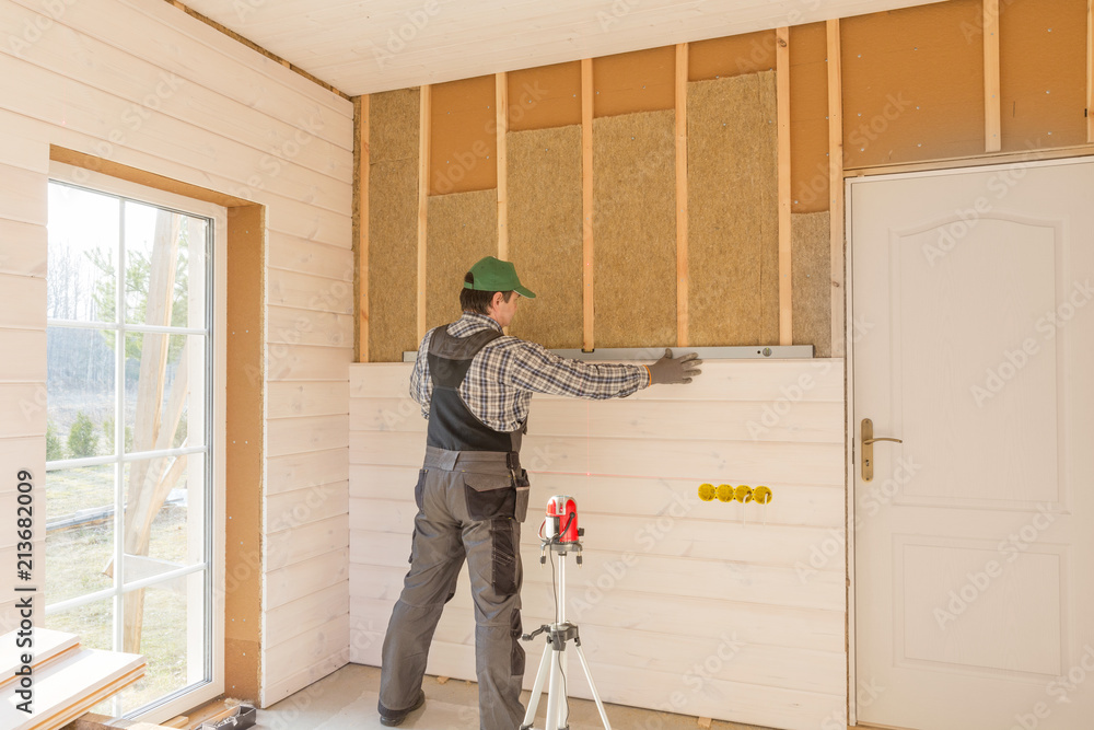 Construction worker thermally insulating eco-wood frame house with wood fiber plates and heat-isolating natural hemp material. Finishing the walls with a white wooden board, using laser line level.