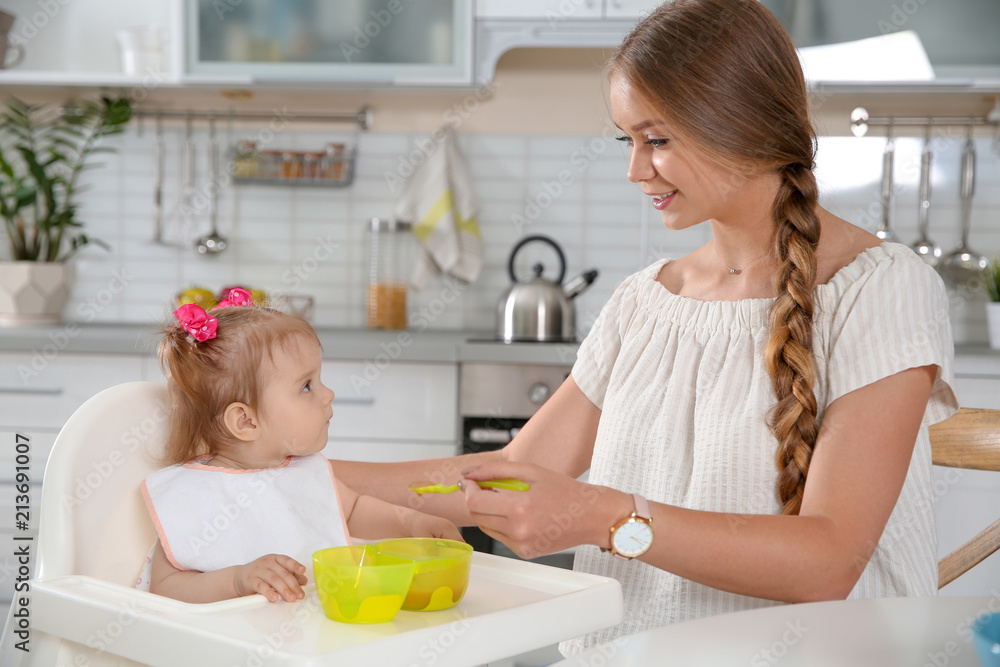 Mother feeding her little baby with healthy food in kitchen