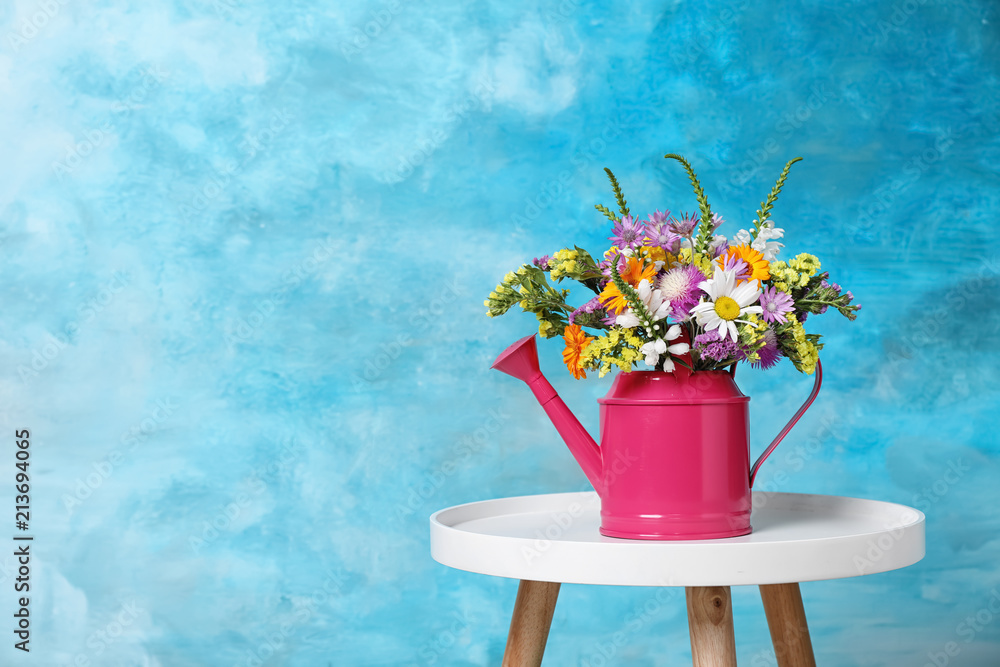 Watering can with beautiful wild flowers on table against color background