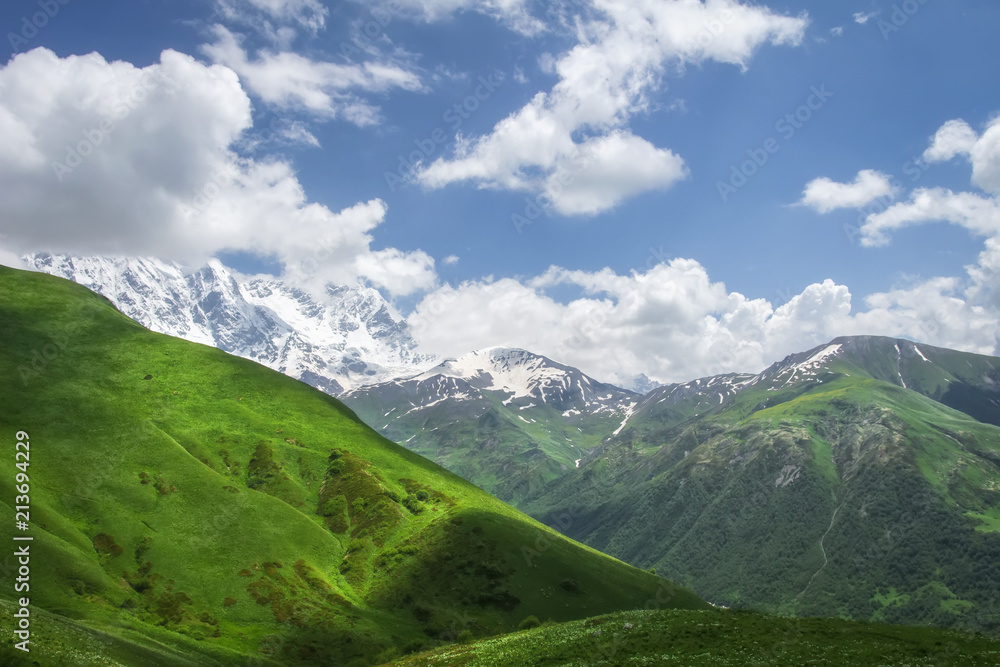 Landscape of mountains and grassy hills on sunny summer day in Svaneti, Georgia. Blue sky and white clouds over georgian mountain. Hills and mounts covered green grass. Scenic georgian wild nature.