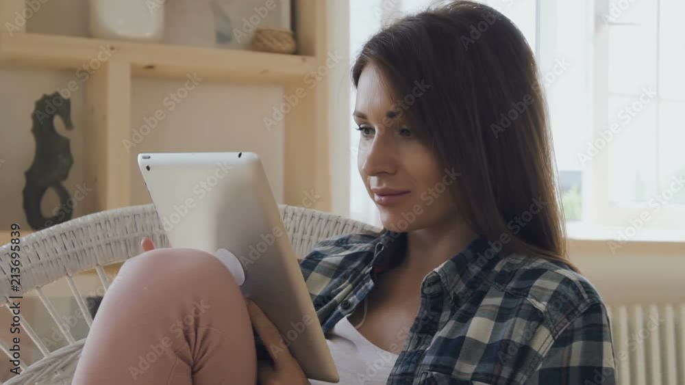 Young woman with brown hair texting on her tablet, smiling, relaxing in white rattan chair at home with cozy interior. Indoors. Portrait. Young woman with brown hair texting on her tablet, smiling, relaxing in white rattan chair at home with cozy interior. Indoors. Portrait.