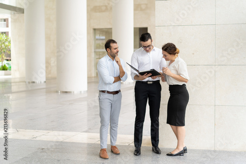 Smiling business team examining documents before meeting in office lobby. Three colleagues, two mid adult Caucasian men and woman discussing papers. Teamwork concept photo