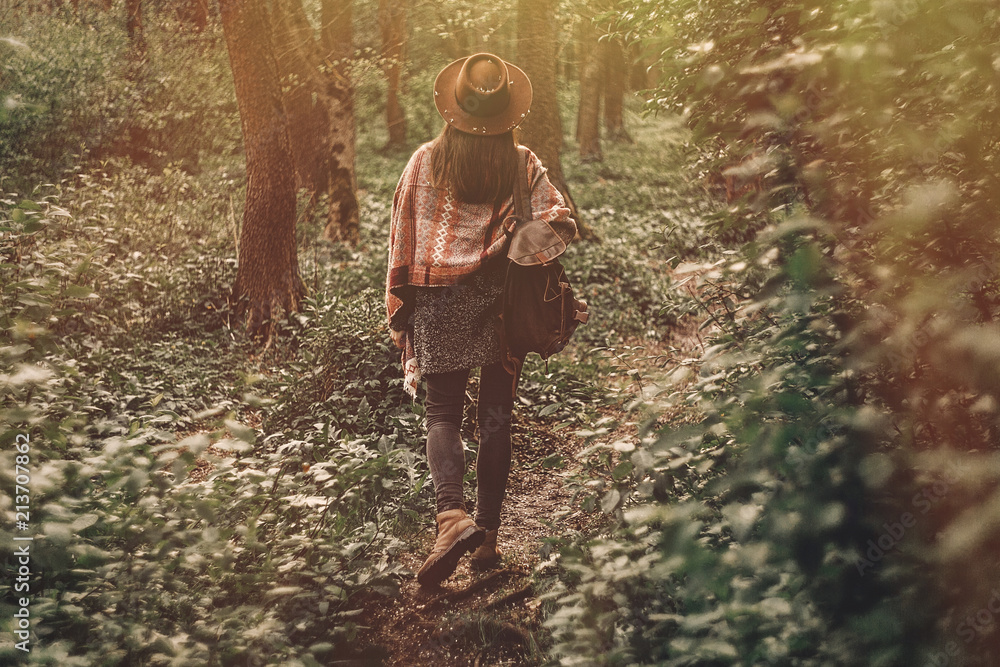 wanderlust concept. stylish hipster girl in hat with backpack walking in woods in evening sunshine. bohemian woman traveler exploring in sunlight. space for text. atmospheric moment