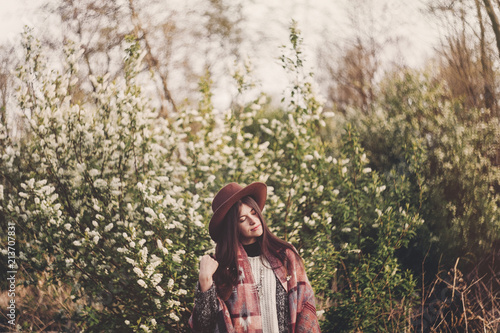 stylish hipster girl in hat and poncho with beautiful hair standing in park in evening sunshine. woman traveler in fashionable outfit relaxing in spring garden. space for text. atmospheric moment
