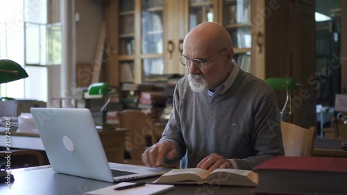 Wallpaper Mural Mature professor working with book and laptop, sitting at table in library, male lecturer doing scientific work using literature and silvery device in reading room with daylight. Concept: academic Torontodigital.ca