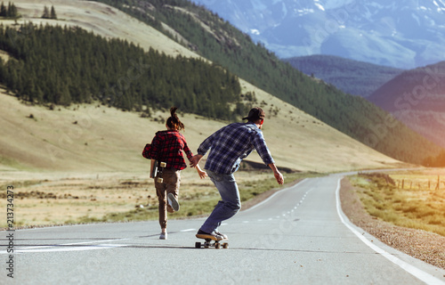 Happy young couple having fun with longboard on the road. Young man and woman skating together on a sunny day in mountain.