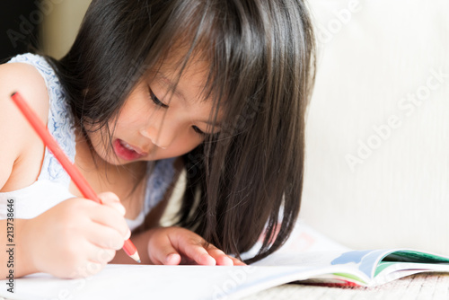 Happy cute little girl smiling and holding red pencil and drawing, writing on a book to do homework.