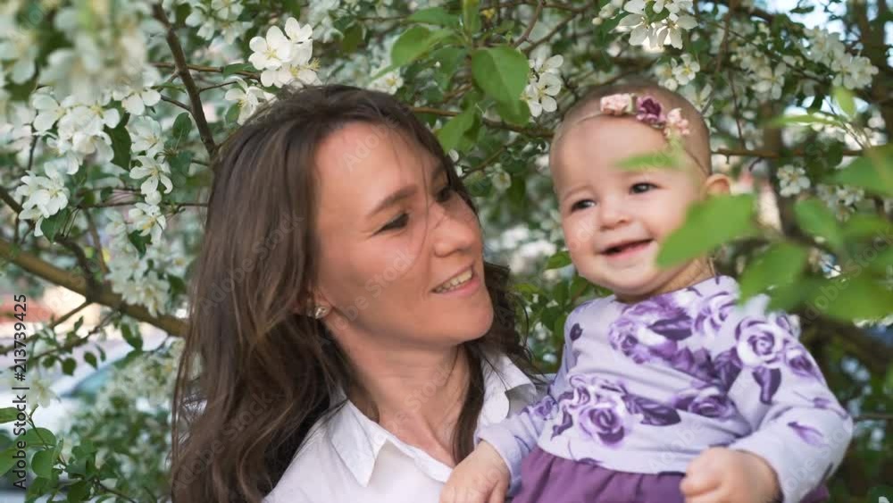 Beautiful active 30s mum play with happy small adorable daughter outside. mother hold, look at her, playing, hug and kiss near with flowers tree at background at summer time in windy day close up Beautiful active 30s mum play with happy small adorable daughter outside. mother hold, look at her, playing, hug and kiss near with flowers tree at background at summer time in windy day close up