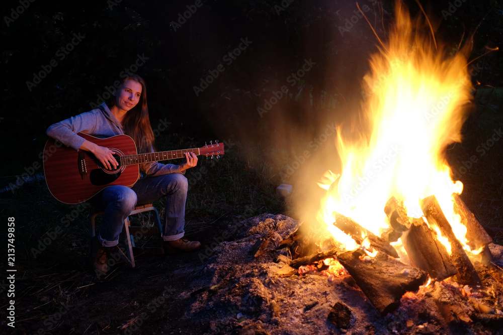 Young redhead woman playing guitar near bonfire at night camp Young redhead woman playing guitar near bonfire at night camp