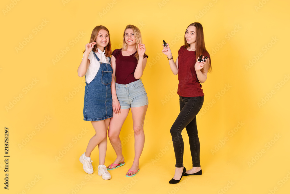 Obraz premium three young girls on a yellow isolated background look directly into the camera. two girls stretch a chewing gum, a third holds the bottles with nail Polish