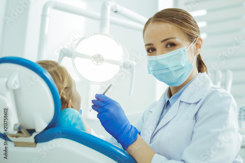 Dental care. Attractive female person covering her face while mask while treating her patient