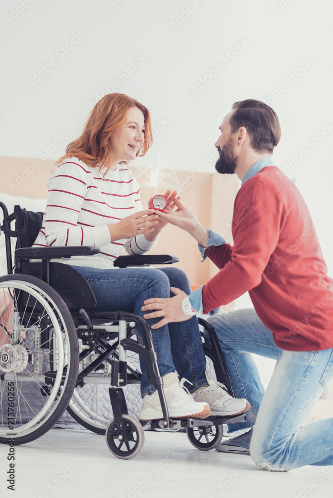 Unexpected proposal. Calm attentive young man standing on one knee and looking at his beloved woman in a wheel chair while asking her to marry him Unexpected proposal. Calm attentive young man standing on one knee and looking at his beloved woman in a wheel chair while asking her to marry him