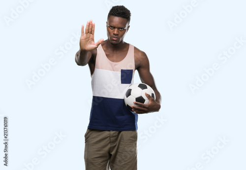 Young african american man holding soccer football ball with open hand doing stop sign with serious and confident expression, defense gesture