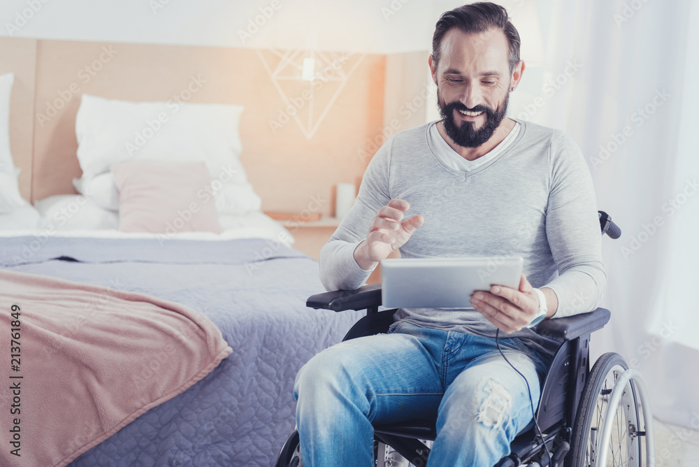 Communicating online. Cheerful friendly young man sitting in a wheelchair and feeling happy while having a pleasant video call