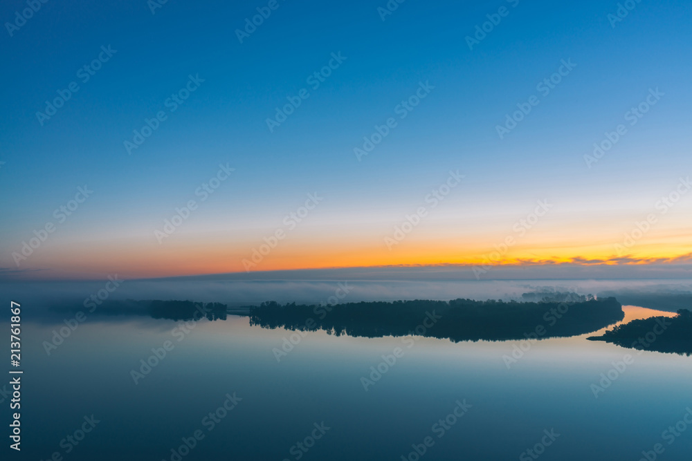 Broad river flows along diagonal shore with forest under thick fog. Early blue sky reflected in water. Yellow glow in picturesque predawn sky. Mystic morning atmospheric landscape of majestic nature