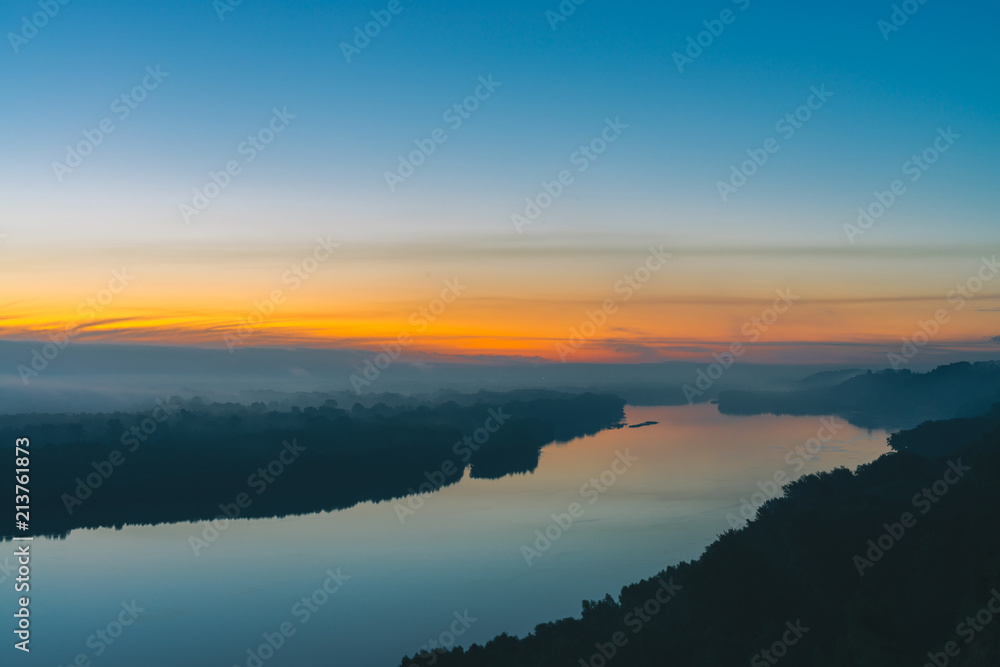 View from high shore on broad river. Riverbank with forest under thick fog. Dawn reflected in water. Yellow glow in picturesque predawn sky. Mystical morning atmospheric landscape of majestic nature.