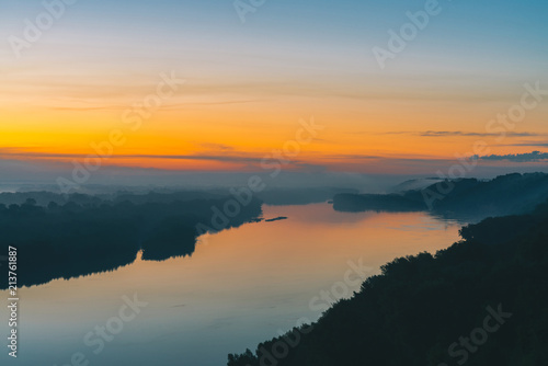 View from high shore on river. Riverbank with forest under thick fog. Gold dawn reflected in water. Yellow glow in picturesque predawn sky. Mystical morning atmospheric landscape of majestic nature.