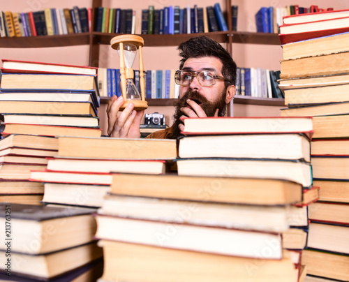 Time flow concept. Teacher or student with beard studying in library. Man, scientist in glasses looks at hourglass. Man on surprised face holds hourglass while studying, bookshelves on background.