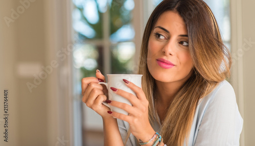 Young beautiful woman smiling and thinking holding a cup of coffee at home