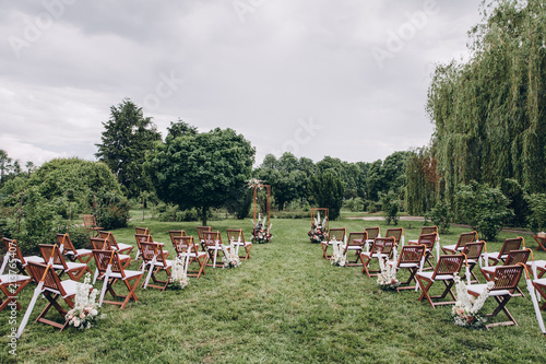 in area of wedding ceremony in park are chairs for guests and arch decorated with flowers