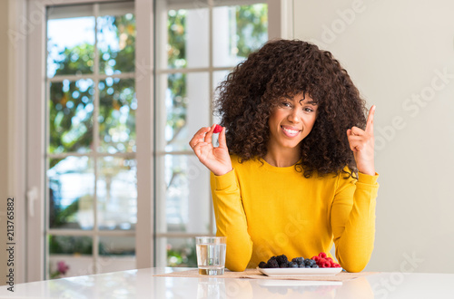 African american woman eating raspberries and blueberries at home surprised with an idea or question pointing finger with happy face, number one