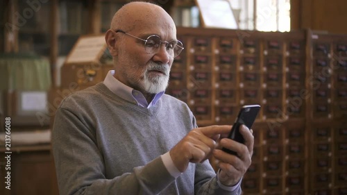 Respectable professor is using smartphone standing in university library, elderly bearded man is looking at phone screen, typing during working day in reading room. Concept: mature person, lifestyle