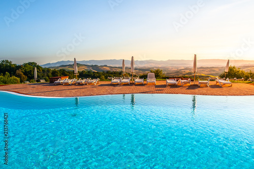 Beautiful luxury swimming pool with bright blue water, umbrellas and sunbeds in Tuscan landscape. Evening summer sunset. Italy.