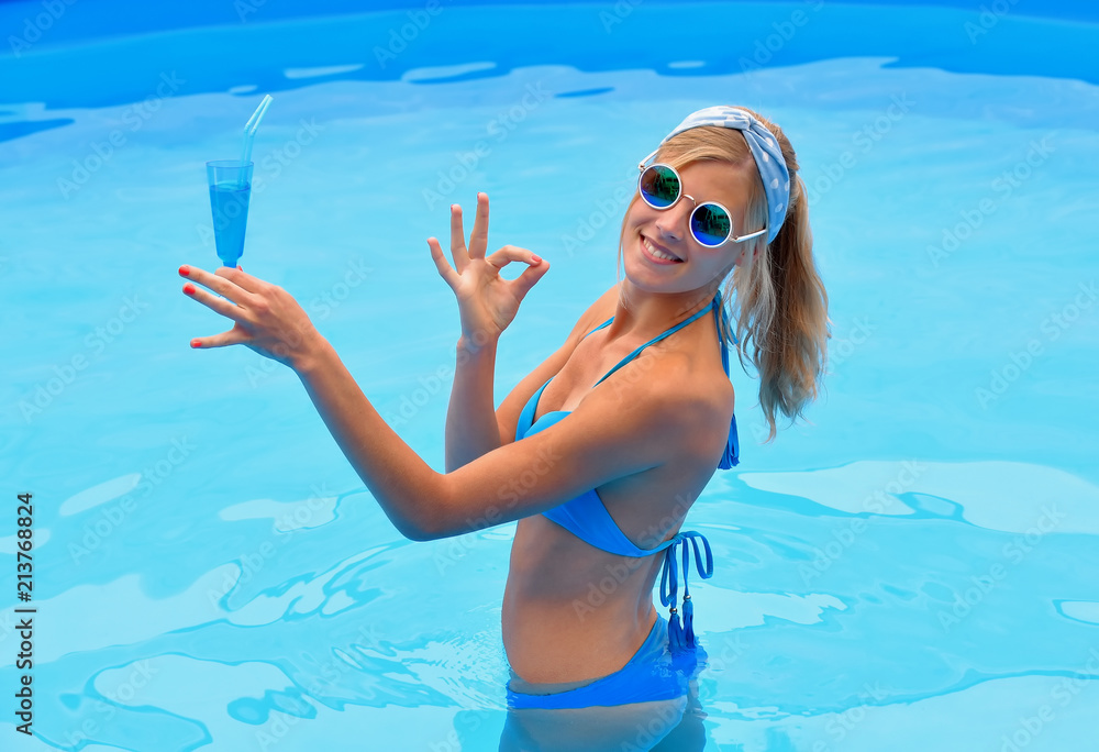 A young girl wears a blue bikini and holds a blue cocktail drink in her hand. She is standing in a pool smiling at the camera.