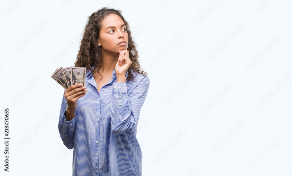 Young hispanic woman holding dollars serious face thinking about question, very confused idea Young hispanic woman holding dollars serious face thinking about question, very confused idea